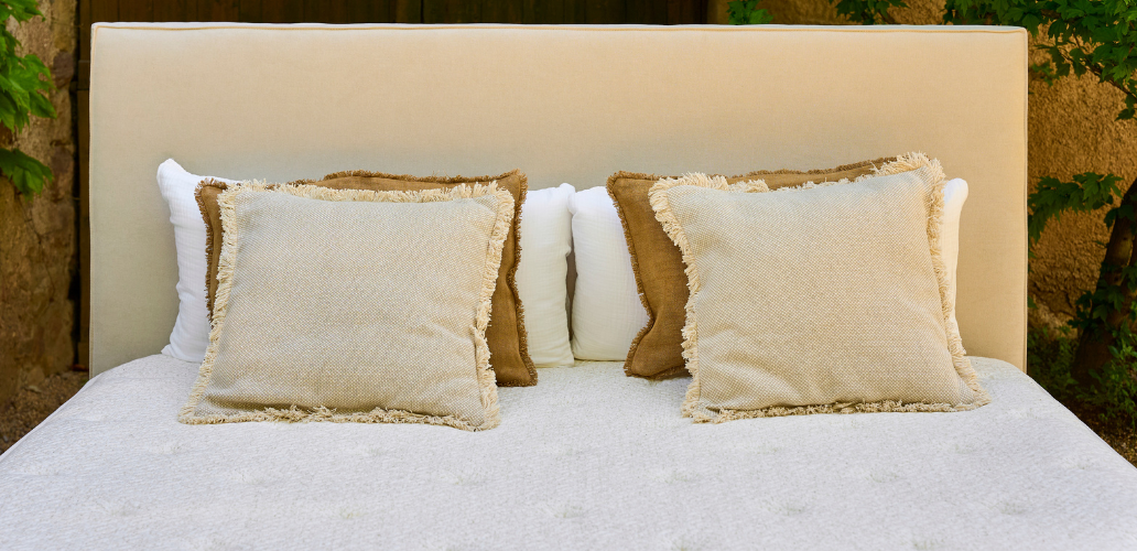 Dormitorio de inspiración mediterránea con cabecero tapizado, colchón de alta calidad y cojines decorativos en tonos naturales, creando un ambiente elegante, acogedor y relajante.